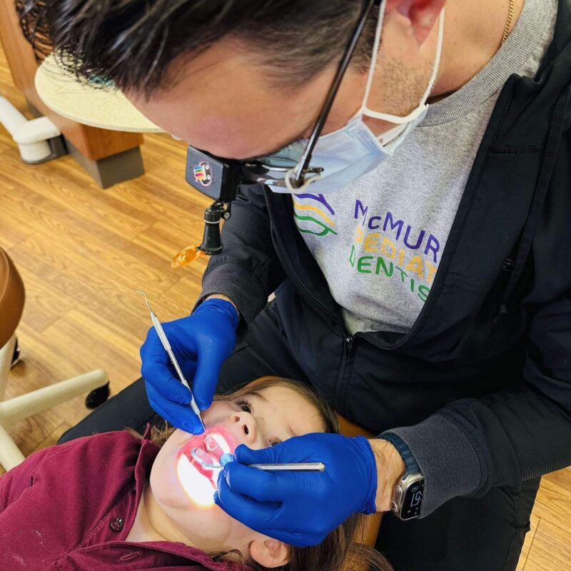 Why Kids Get Cavities Even When Brushing Is Happening, A child smiling while a dentist performs a routine dental exam using a mirror and dental instrument.