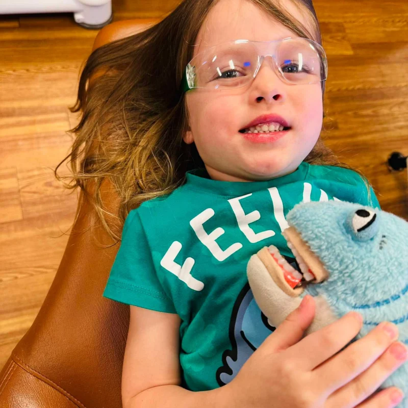 Child smiling in a dental chair while holding a stuffed animal during a pediatric dental visit.