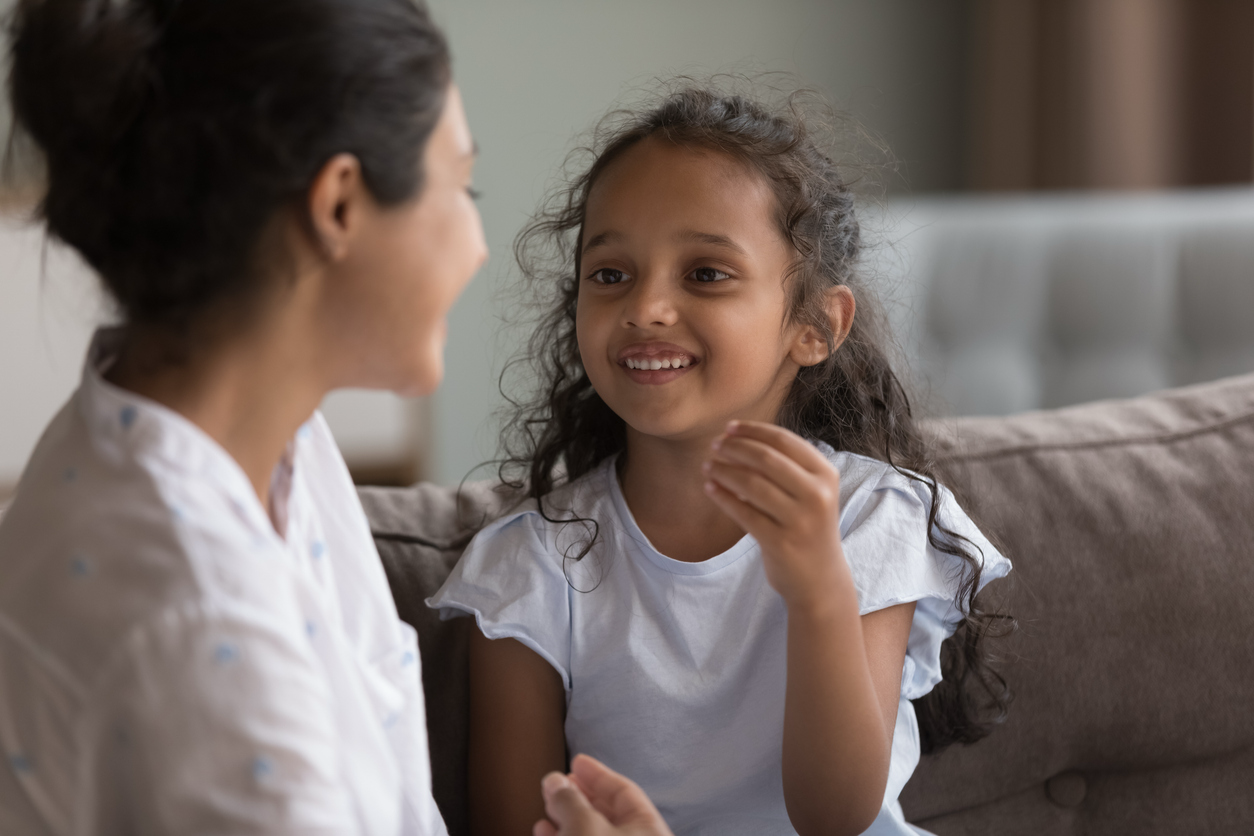 A smiling young girl with curly dark hair talks animatedly with an adult woman in a bright, casual setting.